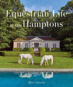 Two white horses graze beside a shimmering pool in front of a charming house nestled in lush greenery—a picturesque moment that echoes the essence of *Equestrian Life in the Hamptons* by Simon & Schuster. On the home’s steps, two people stand, embodying the serene sophistication captured throughout this celebrated equestrian book.
