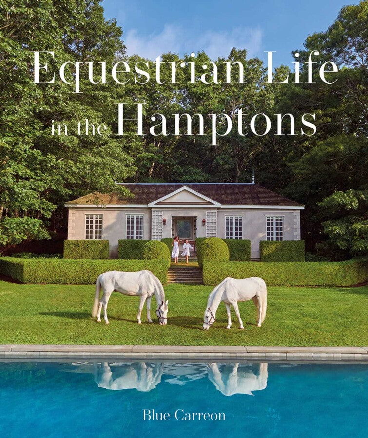 Two white horses graze beside a shimmering pool in front of a charming house nestled in lush greenery—a picturesque moment that echoes the essence of *Equestrian Life in the Hamptons* by Simon & Schuster. On the home’s steps, two people stand, embodying the serene sophistication captured throughout this celebrated equestrian book.