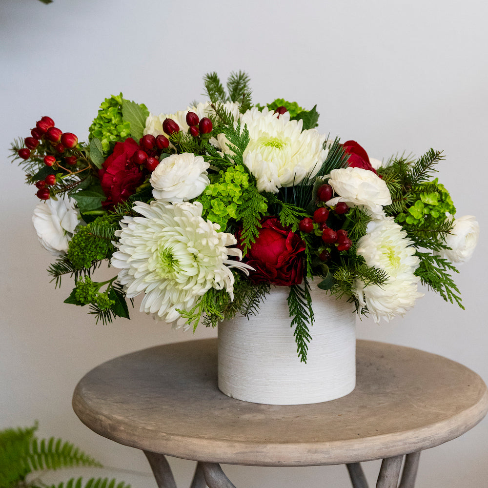 The Hive Floral Studio Live Evergreen Holiday Floral Arrangement features festive blooms and greenery in a white ceramic vase, beautifully displayed on a round wooden table.