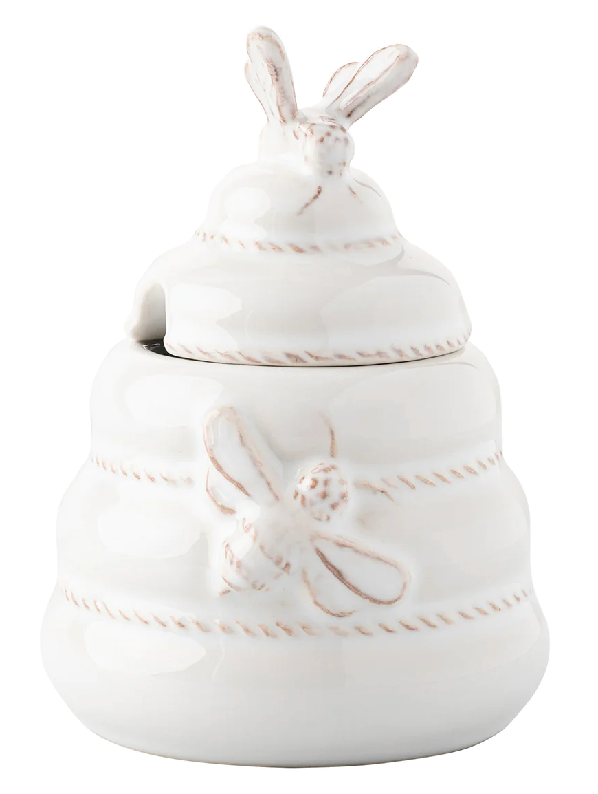 The Juliska Clever Creatures Whitewash Honey Pot is a white ceramic honey jar shaped like a beehive, featuring bee details and a bee-topped lid. It comes complete with a wooden spoon.