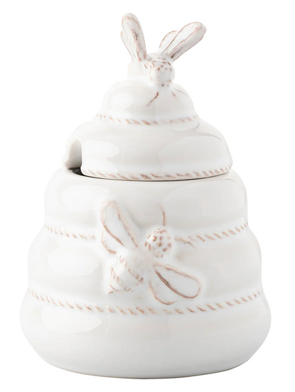 The Juliska Clever Creatures Whitewash Honey Pot is a white ceramic honey jar shaped like a beehive, featuring bee details and a bee-topped lid. It comes complete with a wooden spoon.