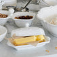 The Juliska Berry & Thread Classic Whitewash Butter Dish rests on a marble countertop, surrounded by white bowls of nuts, dried fruit, flour, and metal measuring cups.