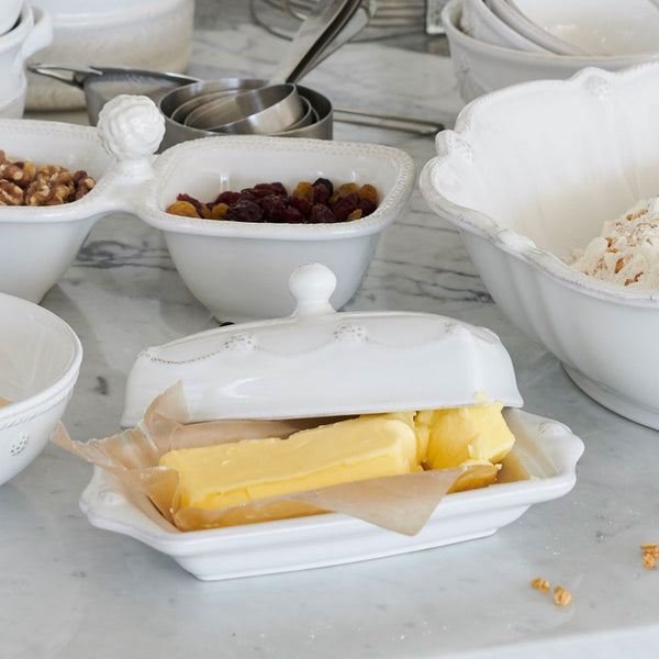 The Juliska Berry & Thread Classic Whitewash Butter Dish rests on a marble countertop, surrounded by white bowls of nuts, dried fruit, flour, and metal measuring cups.