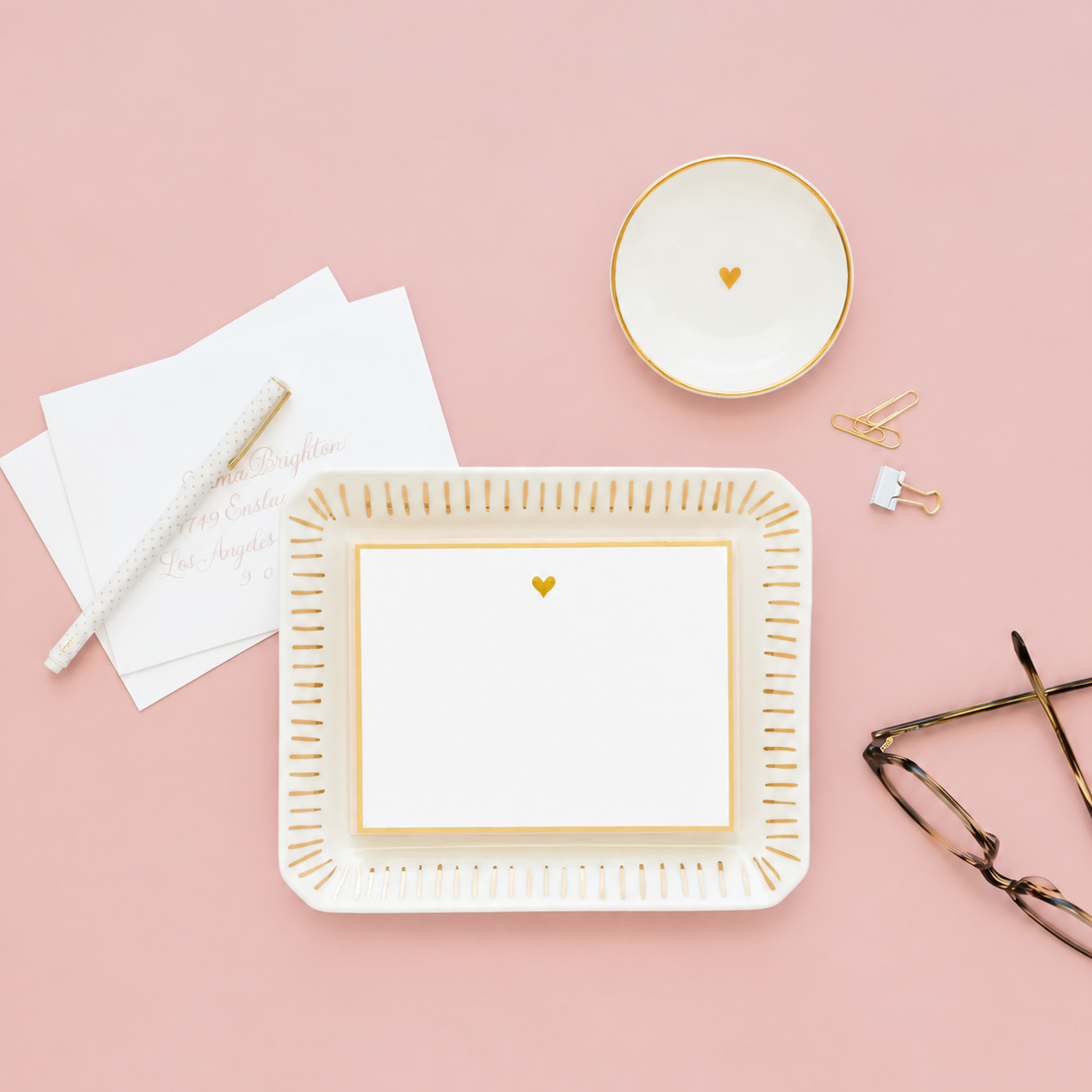 Sugar Paper Small Round Heart Trinket Tray stationery set with elegant gold accents, featuring a ceramic trinket tray, bowl, notecard, envelopes, pen, paper clips, and eyeglasses displayed on a pink background.