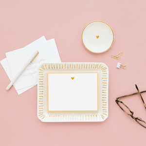 Sugar Paper Small Round Heart Trinket Tray stationery set with elegant gold accents, featuring a ceramic trinket tray, bowl, notecard, envelopes, pen, paper clips, and eyeglasses displayed on a pink background.