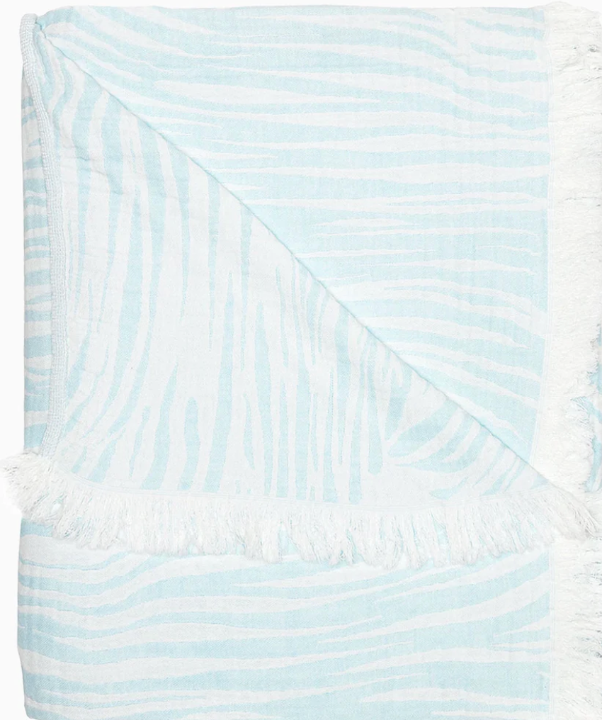 The Daasu Throw Collection by John Robshaw features a light blue and white stonewashed cotton towel with subtle wavy stripes, fringed edges, and a reversible design.