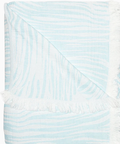 The Daasu Throw Collection by John Robshaw features a light blue and white stonewashed cotton towel with subtle wavy stripes, fringed edges, and a reversible design.