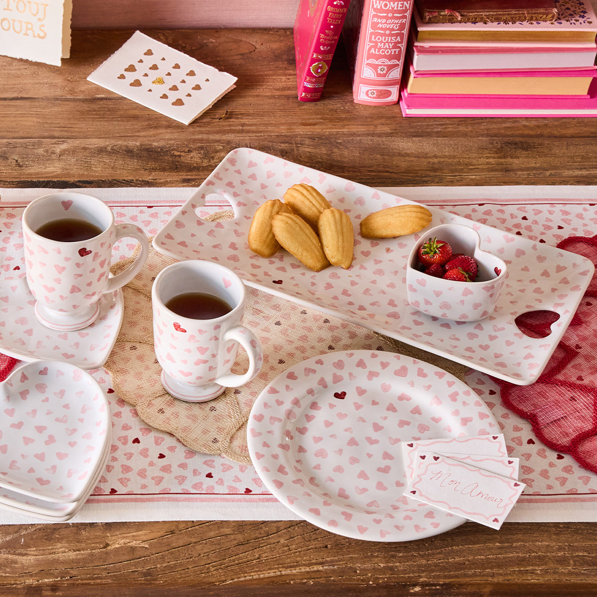 The Juliska Love You More Decor and Tabletop Collection features pink heart plates, two cups with drinks, a rectangular plate with madeleines, strawberries, and name cards on a wooden table with pink-themed accents.