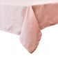A close-up of the Juliska Berry Scallop Tablecloth in light pink linen, elegantly draped over the corner of a rectangular table against a white background.