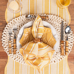 The Juliska Cabana Stripe Tabletop Collection napkin rests in a ring on a white plate, paired with wooden-handled cutlery on a woven placemat. To the top right, a glass of orange liquid adds casual sophistication to the elegant setting.