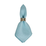 The Berry Scallop Napkin Set by Juliska features light blue woven linen napkins with stitched edges, elegantly folded and secured with a decorative silver napkin ring.