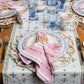 A Juliska Red Villa Stripe Napkin with a scalloped edge folded inside a woven napkin ring, resting on a plate with a blue striped rim and a floral table runner.