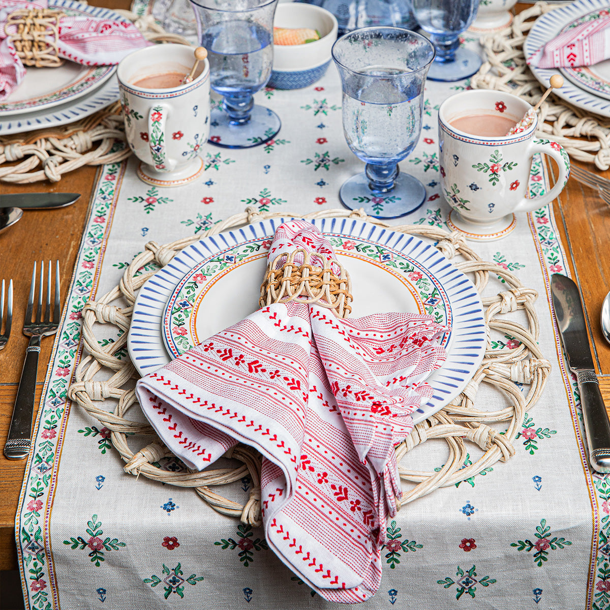 A Juliska Red Villa Stripe Napkin with a scalloped edge folded inside a woven napkin ring, resting on a plate with a blue striped rim and a floral table runner.