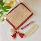 A rectangular Aerin Bordered Raffia Pouch with a red border, two tassels, and a heart-shaped charm, set alongside a glass perfume roller and a light-colored comb.