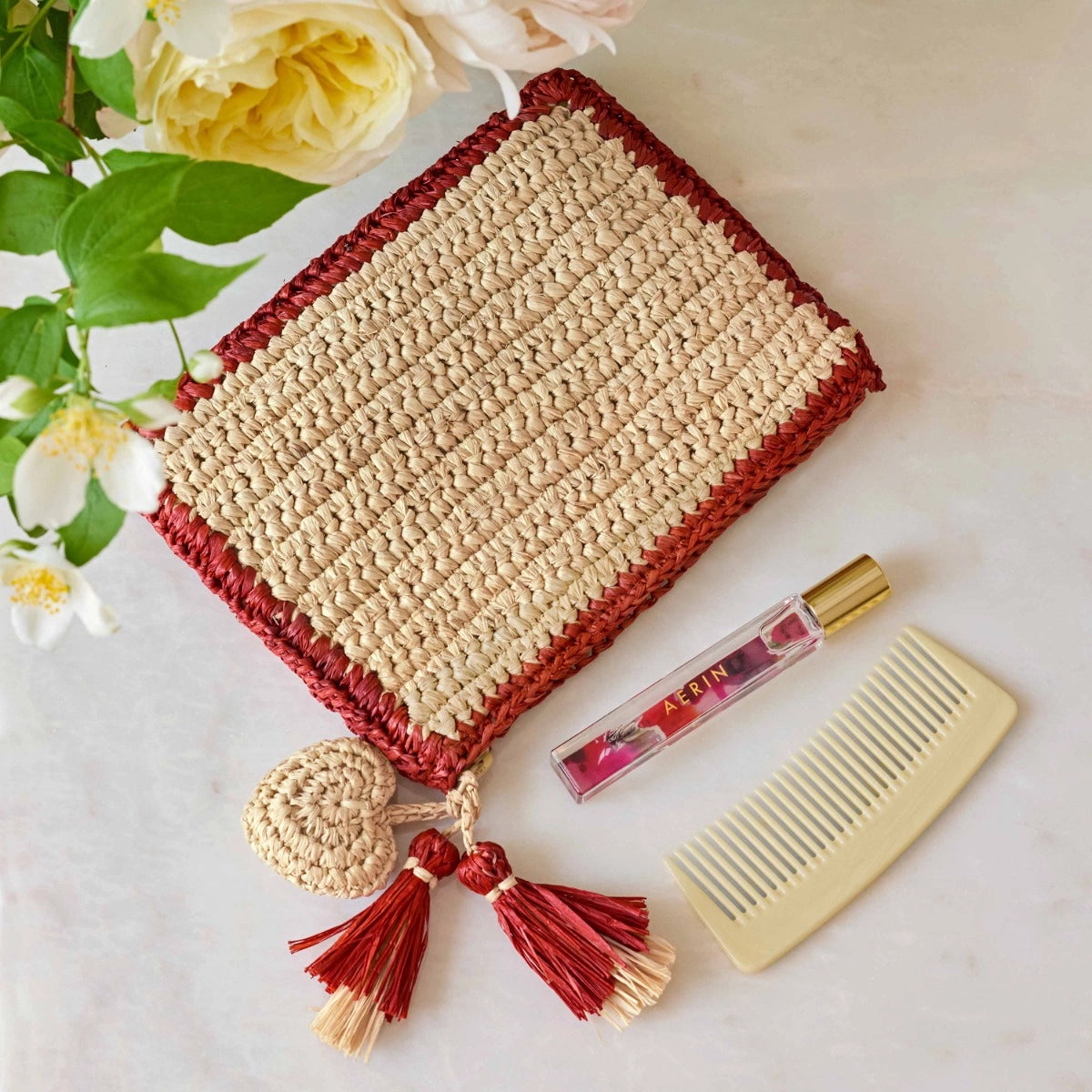 A rectangular Aerin Bordered Raffia Pouch with a red border, two tassels, and a heart-shaped charm, set alongside a glass perfume roller and a light-colored comb.