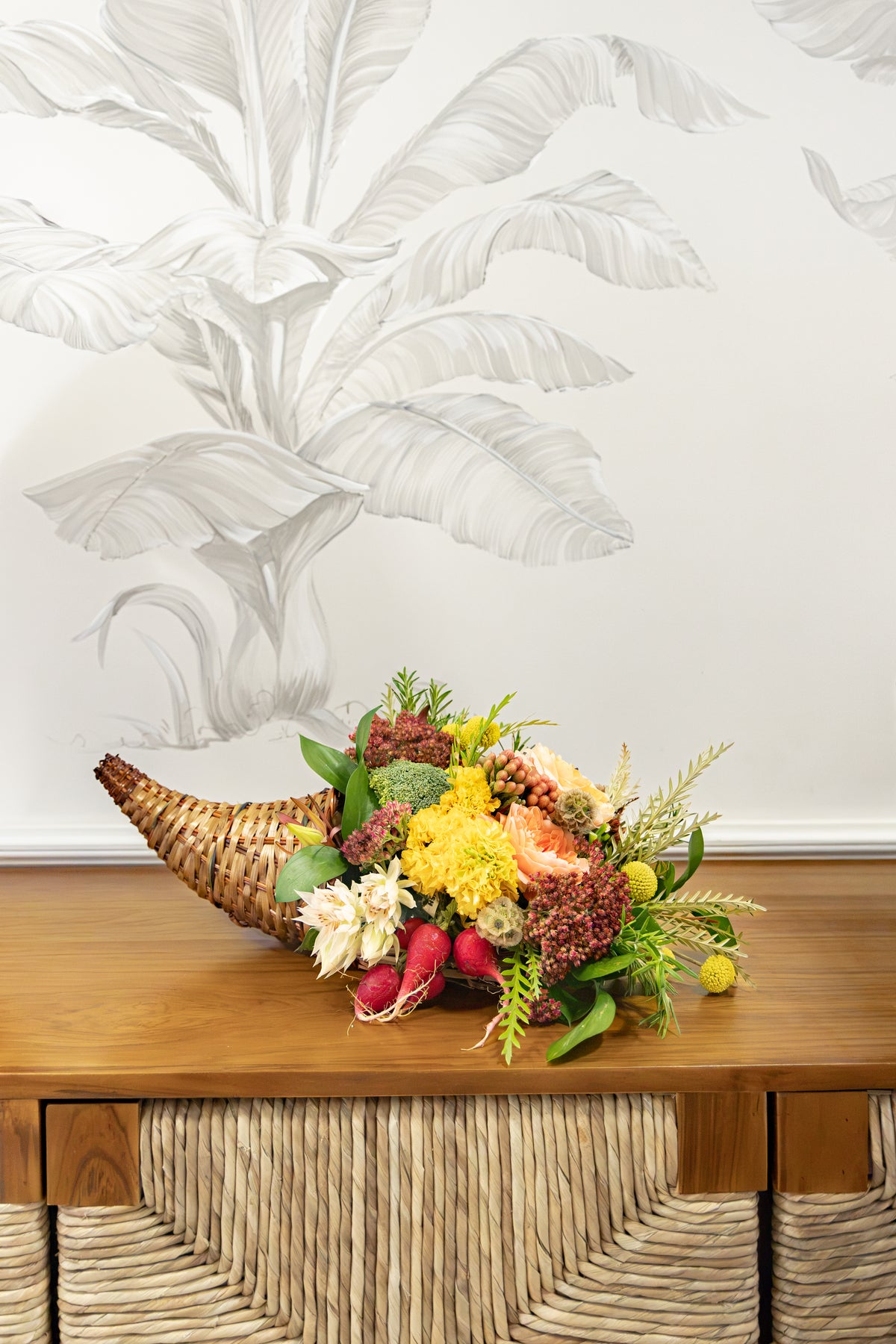 A Hive Floral Class - Thanksgiving Class centerpiece featuring a woven wicker cornucopia filled with yellow chrysanthemums, red radishes, and greenery on a wooden surface.