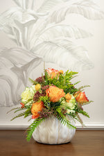 A Hive Floral Class - Thanksgiving Class arrangement of orange and cream roses, greenery, and small purple buds in a white, pumpkin-shaped vase.