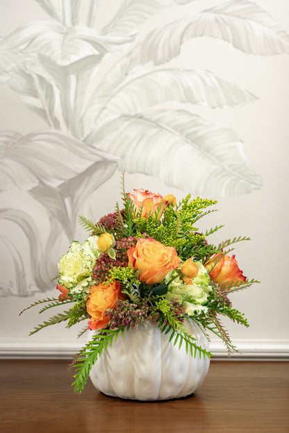 A Hive Floral Class - Thanksgiving Class arrangement of orange and cream roses, greenery, and small purple buds in a white, pumpkin-shaped vase.