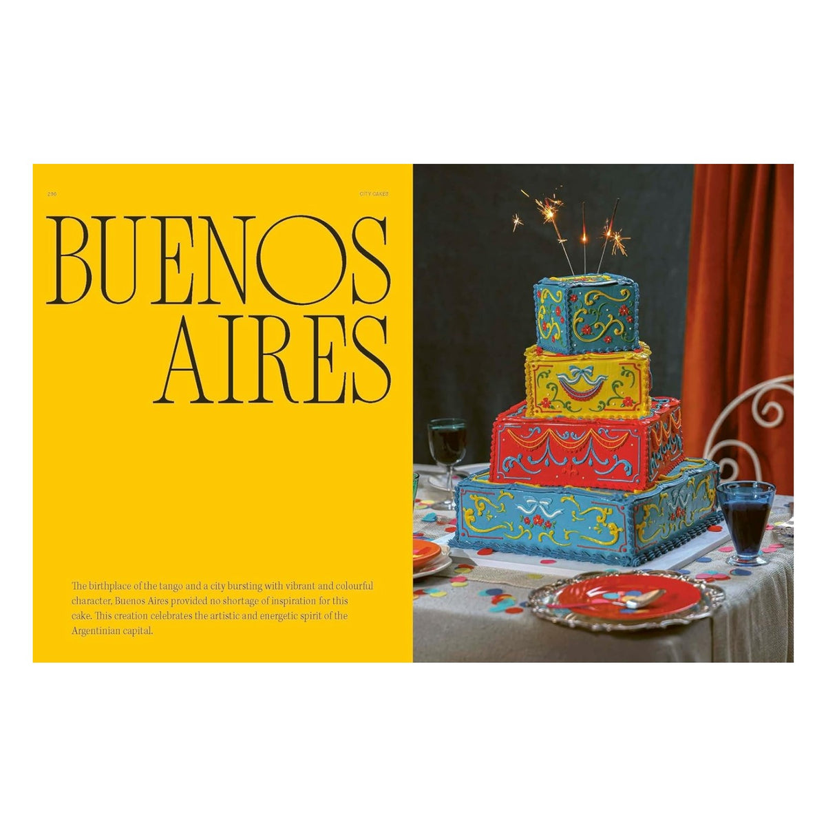 A Fancy Cakes tiered, square-shaped cake with intricate Baroque buttercream piping, decorated in blue, red, and yellow icing with sparklers, displayed on a table with party plates.