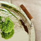 A close-up of the ezrad Bamboo and Stainless Steel Decor Collection tray, featuring a bamboo handle and reflective stainless steel, brings modern elegance with green plant stems and grass-like flowers.