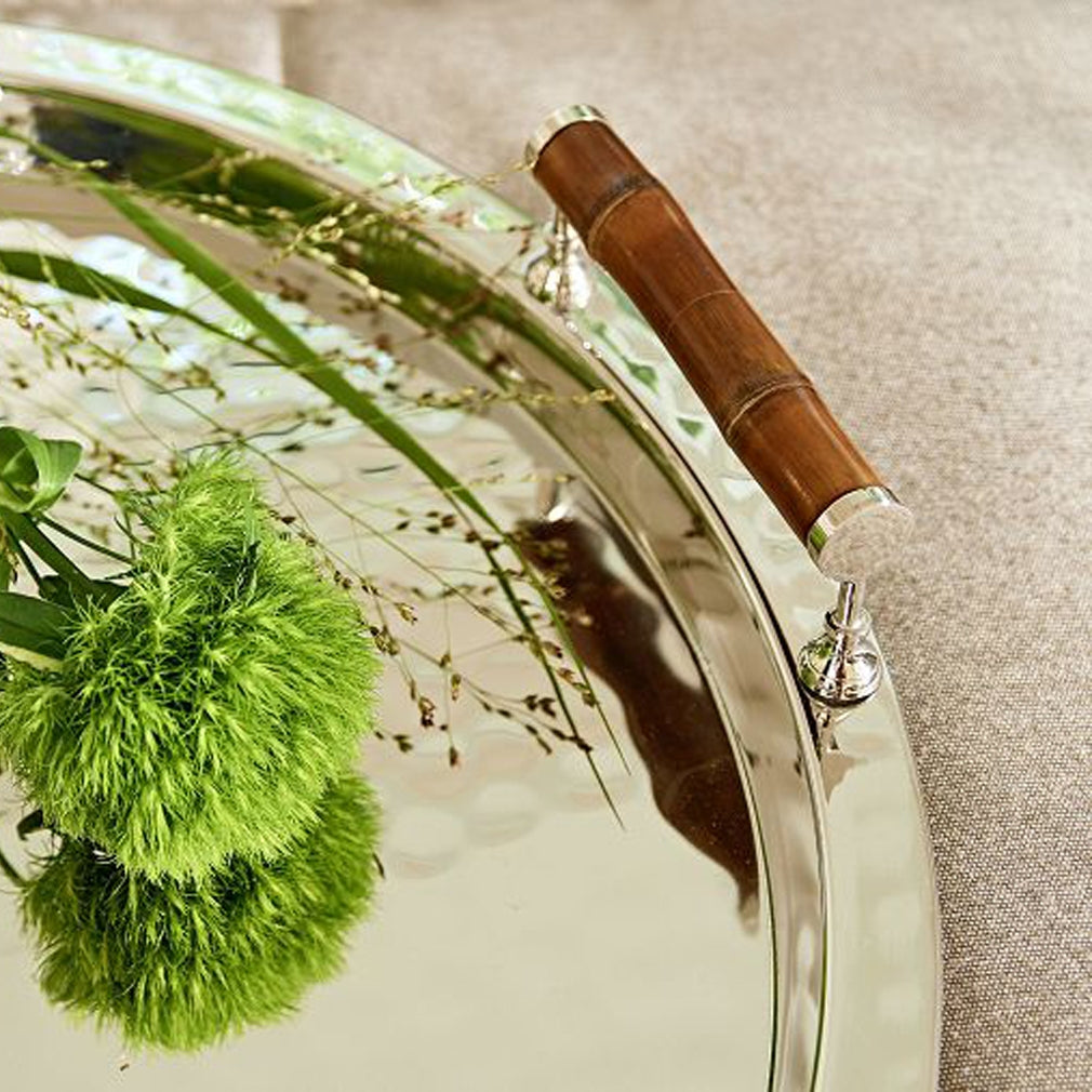 A close-up of the ezrad Bamboo and Stainless Steel Decor Collection tray, featuring a bamboo handle and reflective stainless steel, brings modern elegance with green plant stems and grass-like flowers.
