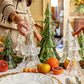 A person arranges Juliska's Berry & Thread Evergreen Tree Collection among fruit, flowers, and a white plate to create festive holiday table decor.