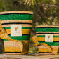 Two Baobab Collection Fety Milalao Candles, in decorative green, black, and yellow striped woven baskets with a gold "Baobab" label and ylang-ylang scent, are placed on a stone surface outdoors against blurred greenery.