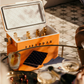 The Fieldbar Gin Trunk, an orange hard case cooler with excellent thermal performance, sits on patterned tile filled with ice and bottled drinks, surrounded by glasses and a tray of cherries.