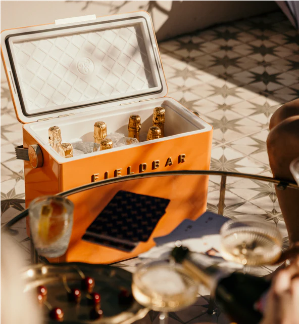 The Fieldbar Gin Trunk, an orange hard case cooler with excellent thermal performance, sits on patterned tile filled with ice and bottled drinks, surrounded by glasses and a tray of cherries.