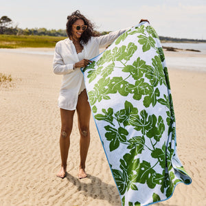 On a sandy beach, a woman holds an oversized Weezie x Peter Dunham Fig Beach Towel with green leaf patterns. She wears a white shirt and sunglasses, enjoying the luxury as serene water and distant trees form the backdrop.