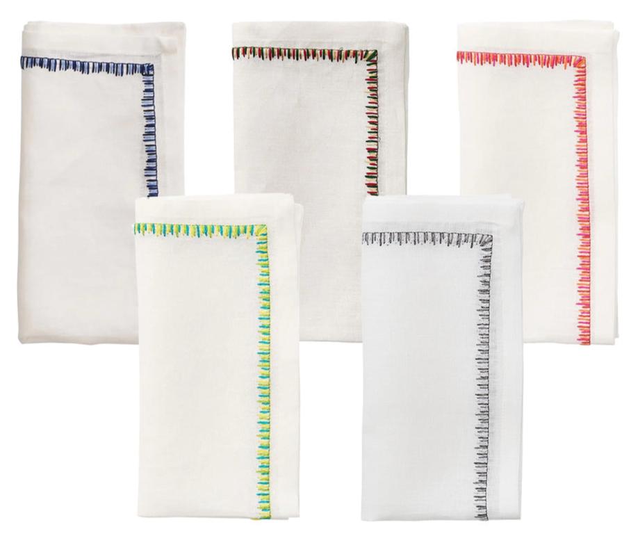 The Filament Napkin Set by Kim Seybert displays five white linen napkins, each with unique embroidered stitching—blue, red, green, black, and multicolored—shown against a white background.