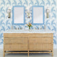 A double-sink vanity with wicker drawers, wall-mounted lights, blue patterned wallpaper, and Fleur Home's Garden District Coquette Rectangle Mirror in Denim Wash evoke New Orleans charm. A small vase with flowers sits between the sinks.