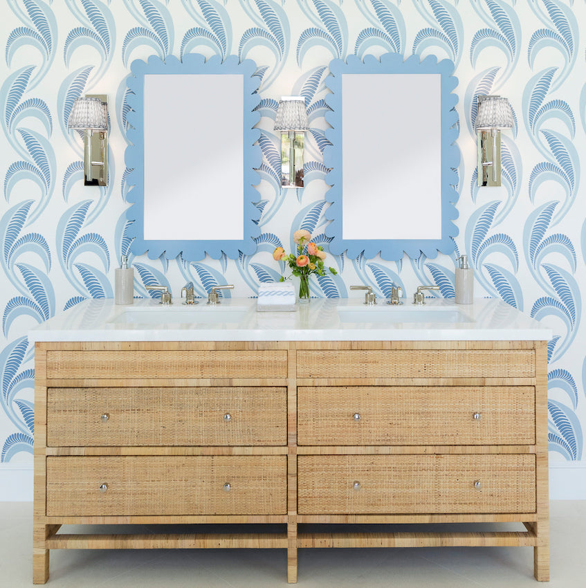 A double-sink vanity with wicker drawers, wall-mounted lights, blue patterned wallpaper, and Fleur Home's Garden District Coquette Rectangle Mirror in Denim Wash evoke New Orleans charm. A small vase with flowers sits between the sinks.