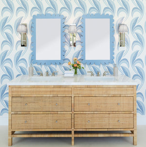 A double-sink vanity with wicker drawers, wall-mounted lights, blue patterned wallpaper, and Fleur Home's Garden District Coquette Rectangle Mirror in Denim Wash evoke New Orleans charm. A small vase with flowers sits between the sinks.
