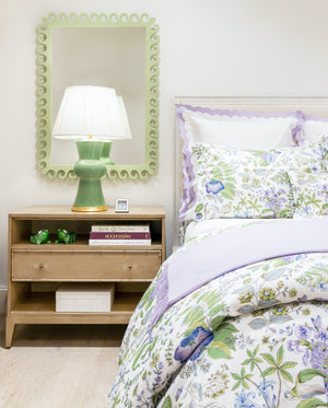 A neatly made bed with floral bedding stands next to a wooden nightstand adorned with an Hwang Bishop Ginger Table Lamp, books, and a framed mirror with a decorative scalloped edge.