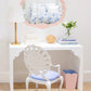 A white vanity table with the Fleur Home Carnival Proteus Mirror, a white lamp, decorative chair, books, flowers, and a pink wastebasket brighten this neatly arranged New Orleans-inspired room.