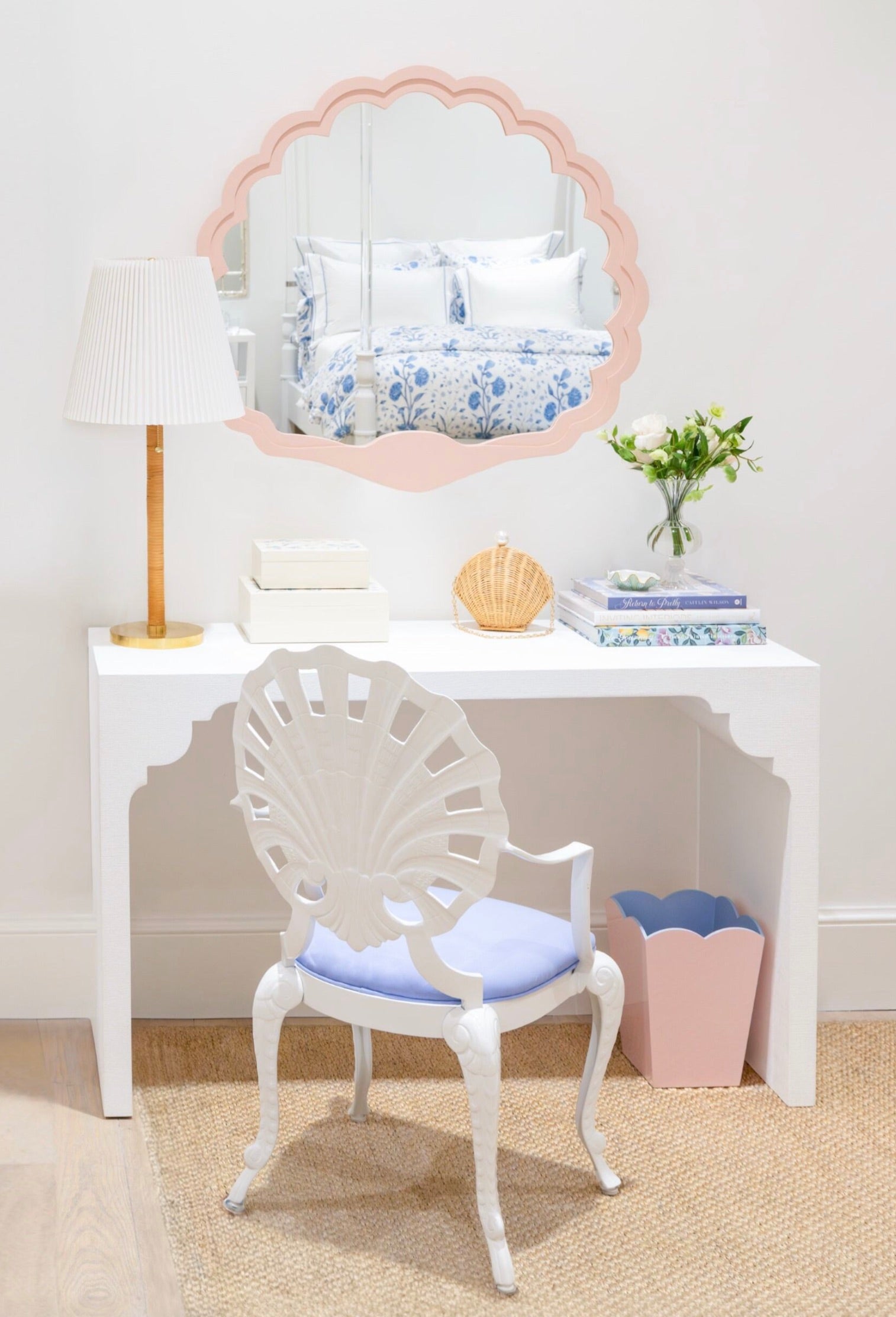 A white vanity table with the Fleur Home Carnival Proteus Mirror, a white lamp, decorative chair, books, flowers, and a pink wastebasket brighten this neatly arranged New Orleans-inspired room.