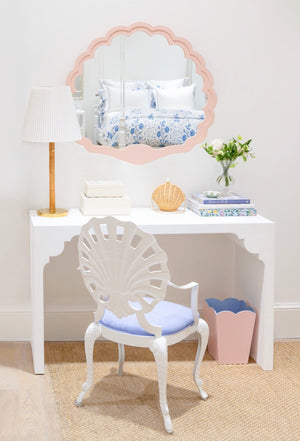 A white vanity table with the Fleur Home Carnival Proteus Mirror, a white lamp, decorative chair, books, flowers, and a pink wastebasket brighten this neatly arranged New Orleans-inspired room.