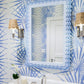 A bathroom features the Carnival Krewe Rectangle Mirror by Fleur Home in pastel blue with scalloped edges, patterned blue wallpaper, silver faucets, wicker-wrapped wall sconces, and a white orchid on the counter.