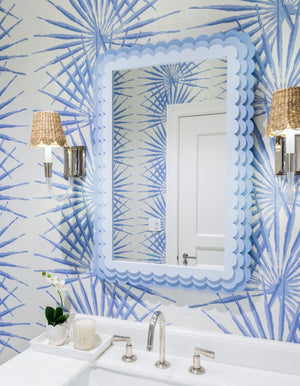 A bathroom features the Carnival Krewe Rectangle Mirror by Fleur Home in pastel blue with scalloped edges, patterned blue wallpaper, silver faucets, wicker-wrapped wall sconces, and a white orchid on the counter.