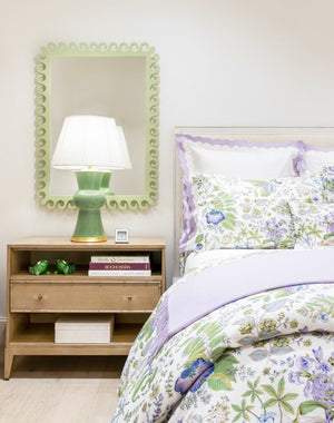 A bedside table with books, a Cooking Apple Green lamp, and a small photo sits next to a bed with floral bedding and a Fleur Home Carnival Gras Rectangle Mirror hanging above.