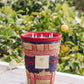 A colorful woven basket with a label reading "Baobab Collection Frida Draozy Frida Candle" from Baobab placed on a wooden surface, surrounded by greenery and lemon trees, evoking the surrealism of a dreamlike garden.