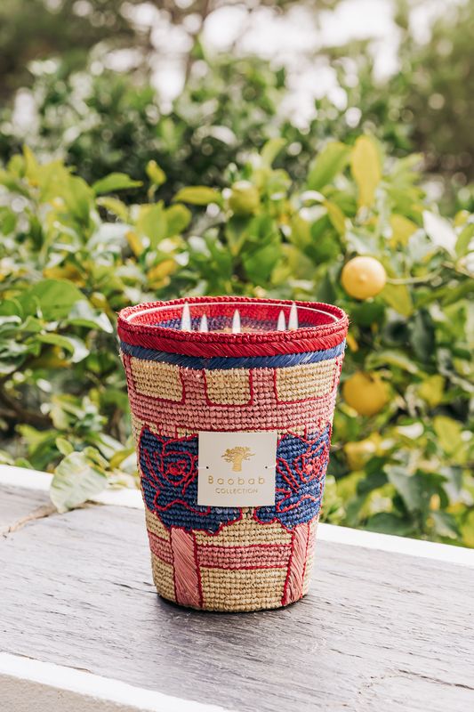 A colorful woven basket with a label reading "Baobab Collection Frida Draozy Frida Candle" from Baobab placed on a wooden surface, surrounded by greenery and lemon trees, evoking the surrealism of a dreamlike garden.