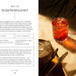 A glass of Negroni Sbagliato—served over ice with a twist of orange peel—rests on a sunlit table beside a white napkin and metal tongs, making it an ideal recipe from Simon & Schuster’s Friday Night Cocktails for anyone seeking delicious homemade mixed drinks.