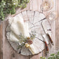 A rustic table features Kim Seybert's Fringe Napkin Set with festive greenery, a wood slice plate, knife, fork, two glasses, artificial snow, and lurex-accented greenery on a wooden surface.