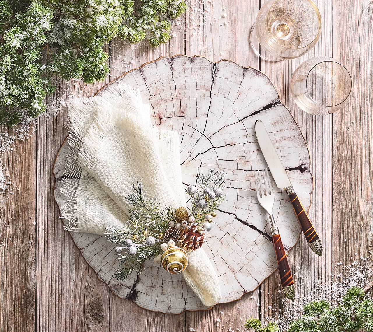 A rustic table features Kim Seybert's Fringe Napkin Set with festive greenery, a wood slice plate, knife, fork, two glasses, artificial snow, and lurex-accented greenery on a wooden surface.