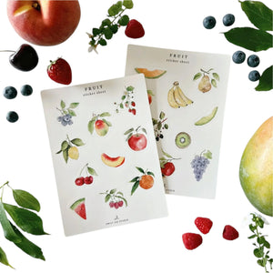 Two Emily Lex Studio Sticker Sheet Sets featuring fruit themes are surrounded by assorted real fruits and green leaves arranged on a white background.