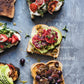 Four slices of toast topped with an array of fruits, vegetables, and spreads are artfully arranged on a gray surface beside the word “Breakfast”—a cozy and inviting scene inspired by recipes from the Half Baked Harvest Cookbook by Penguin Random House.