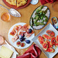 A table set in true Suzanne Lenzer fashion features a grazing assortment inspired by *Graze* from Penguin Random House, with sliced pizza, grilled peppers, prosciutto with figs, tomato bread, cheese, canned fish, drinks, and red napkins arranged on a wooden surface for an inviting tapas-style spread.