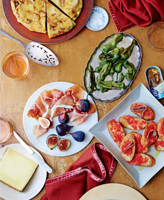 A table set in true Suzanne Lenzer fashion features a grazing assortment inspired by *Graze* from Penguin Random House, with sliced pizza, grilled peppers, prosciutto with figs, tomato bread, cheese, canned fish, drinks, and red napkins arranged on a wooden surface for an inviting tapas-style spread.